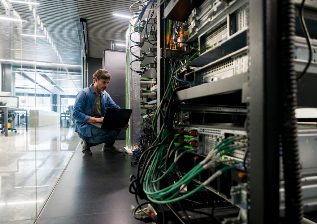 IT technician managing network hardware in an office server room, illustrating the physical risks and lifecycle of assets in cloud-first environments