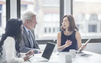 Business meeting with diverse professionals discussing IT asset disposition strategies, featuring a woman presenting with a tablet, a man listening attentively, and another woman taking notes.