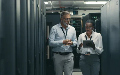 Professionals discussing IT asset management in a data centre, reviewing tablet data amidst server racks.